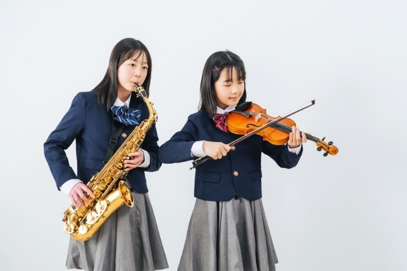 Two school-aged girls in matching school uniforms perform music, with one playing a saxophone and the other playing a violin.