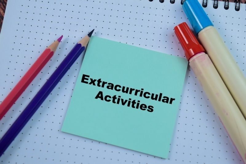 A blue note card labeled “Extracurricular Activities” sits on a dotted notebook, surrounded by colorful pencils and markers.