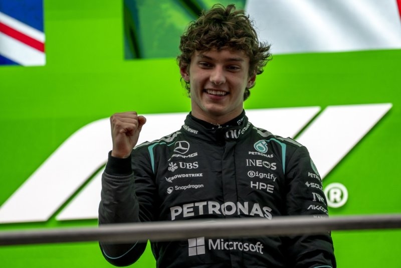 Formula 1 driver in a black racing suit celebrating with a raised fist in front of a green F1 backdrop.