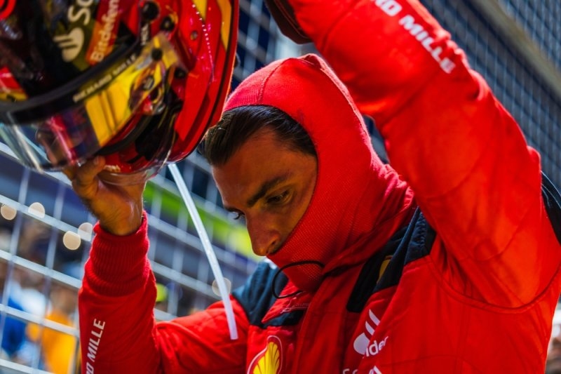 Formula 1 driver in a red racing suit and helmet preparing for a race inside a pit or paddock area.