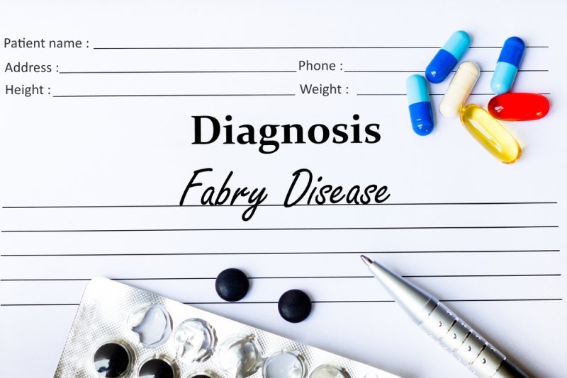 Medical diagnosis form labeled “Fabry Disease,” surrounded by colorful capsules, loose tablets, a blister pack, and a pen on a white surface.