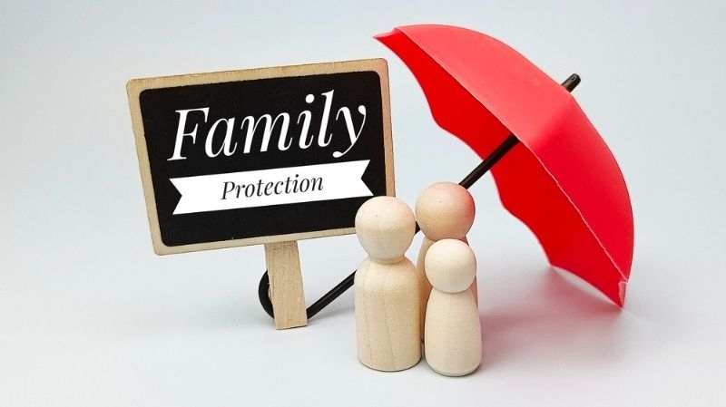 Wooden family figures standing under a red umbrella beside a ‘Family Protection’ sign.