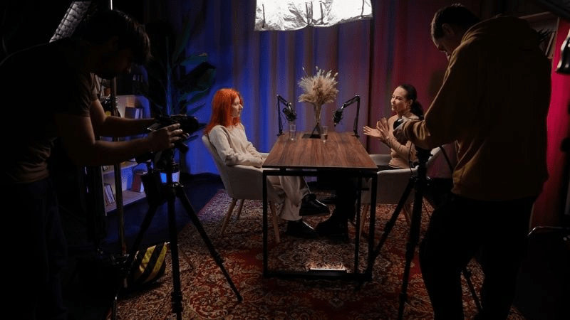 Podcast recording setup with two women speaking at a table while camera crew films in a studio.