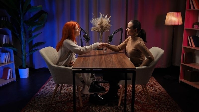 Two women shaking hands across a podcast table with microphones in a softly lit recording studio.
