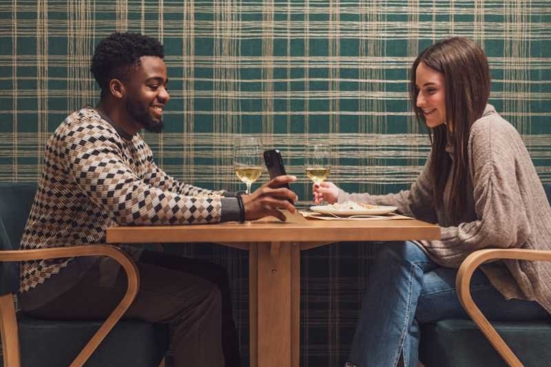 A couple sitting together at a restaurant, smiling and talking while looking at a phone.