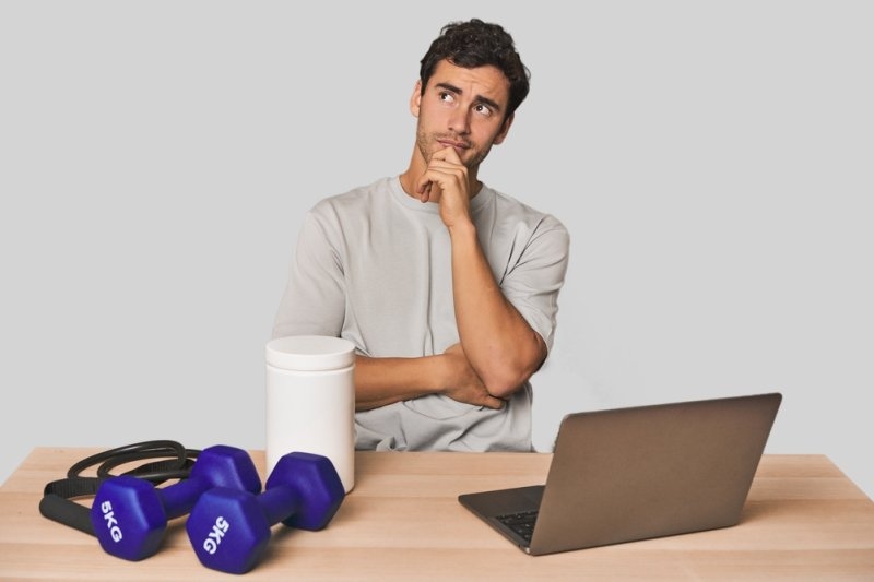 A man sits at a table with dumbbells, a supplement container, and a laptop, appearing to consider a fitness-related business or decision.