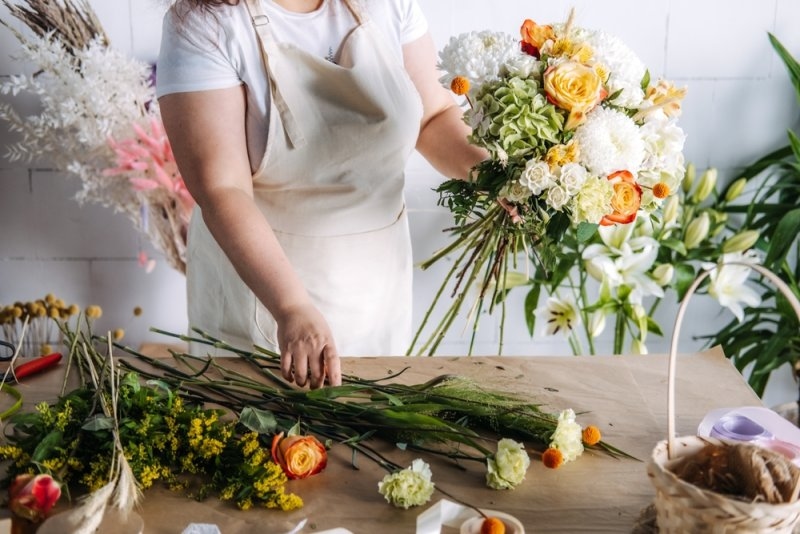 Florist creating a large wedding bouquet with orange and white blooms on a wooden table. 