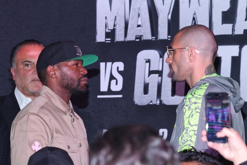 Floyd Mayweather Jr. wearing a beige jacket and black cap during a boxing press event, facing another man against a promotional backdrop.