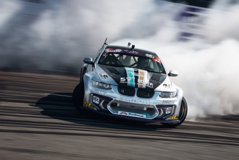Modified BMW drift car sliding around a racetrack with smoke billowing from its tires.