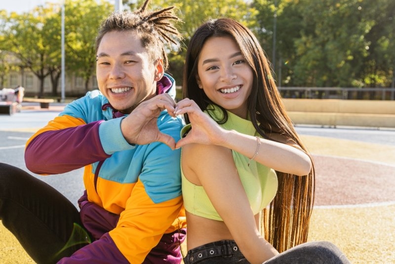 Smiling couple sitting outdoors and forming a heart shape with their hands.