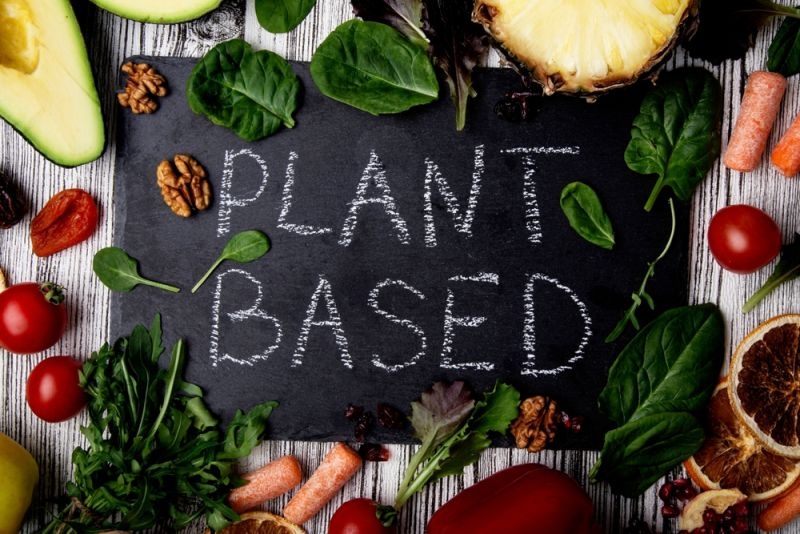 Chalkboard labeled “PLANT BASED” surrounded by leafy greens, vegetables, fruits, nuts, and other plant-based foods.
