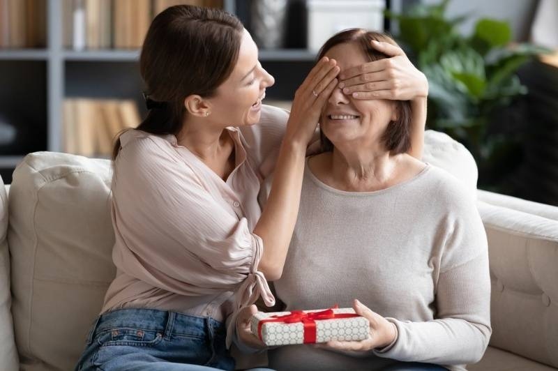 Woman surprising an older woman with a gift box while playfully covering her eyes on a couch.