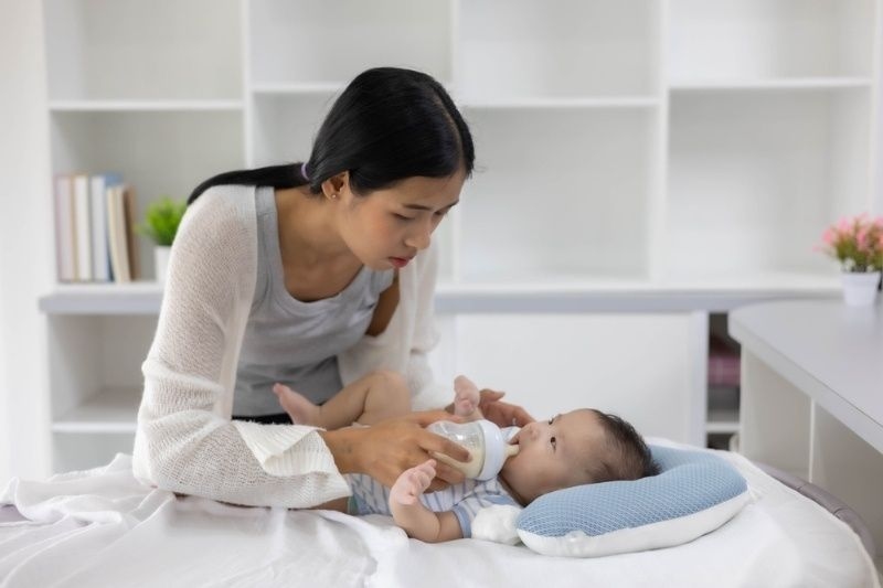 Mother feeding a baby with a bottle while the baby lies on a pillow in a bright home setting.