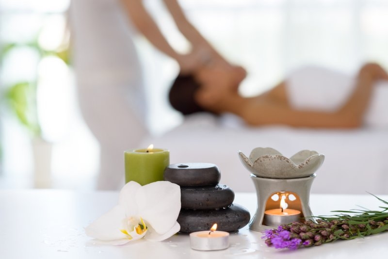 A woman holding a lit candle in a relaxing bedroom setting, surrounded by spa-inspired elements, suggesting a self-care or wellness gift.