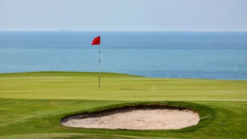 Scenic coastal golf course with a neatly maintained green, a sand bunker, and a red flag marking the hole overlooking the ocean.