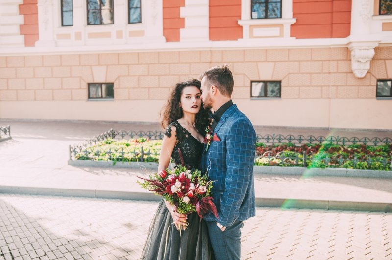 Couple posing outdoors, with a woman wearing a black gothic dress and holding a bouquet beside a man in a blue suit.