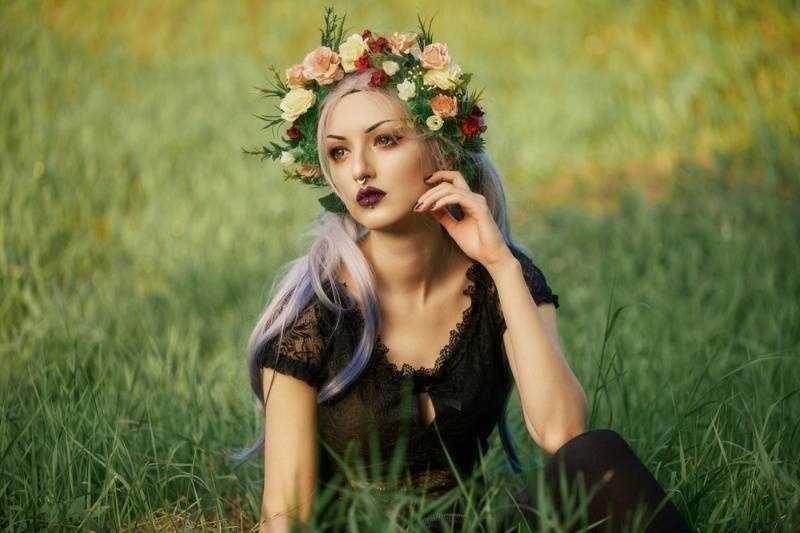 A woman with colorful hair and a floral crown sits in a grassy field, wearing a black lace outfit and dramatic makeup with a dark, alternative aesthetic.