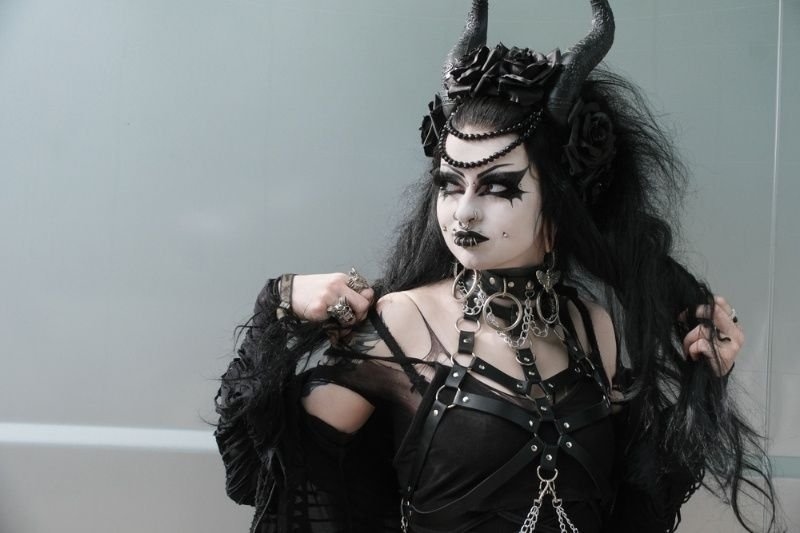 A woman poses in an elaborate gothic outfit featuring black clothing, dramatic makeup, dark accessories, long black hair, and a horned headpiece.