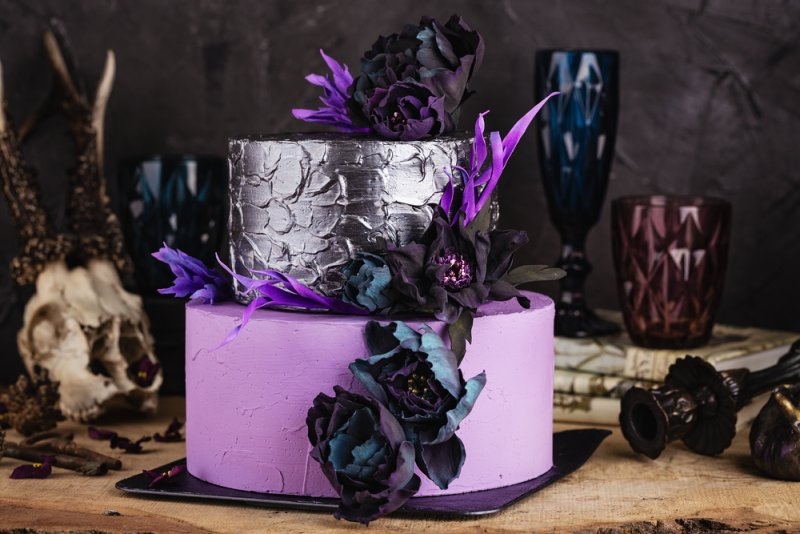 A dramatic two-tier purple and silver wedding cake decorated with dark flowers against a gothic-themed backdrop.