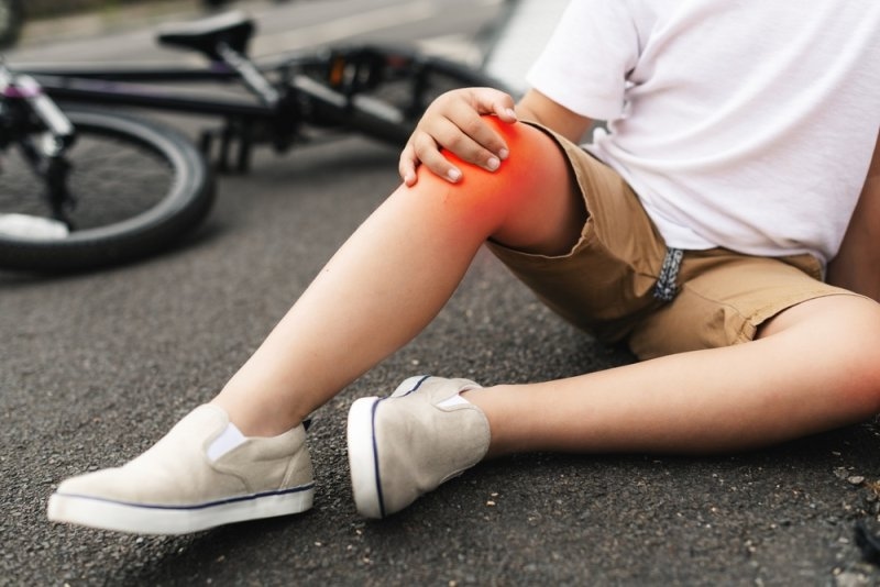 Child sitting on pavement with a scraped knee, highlighted in red. A bicycle lies nearby.