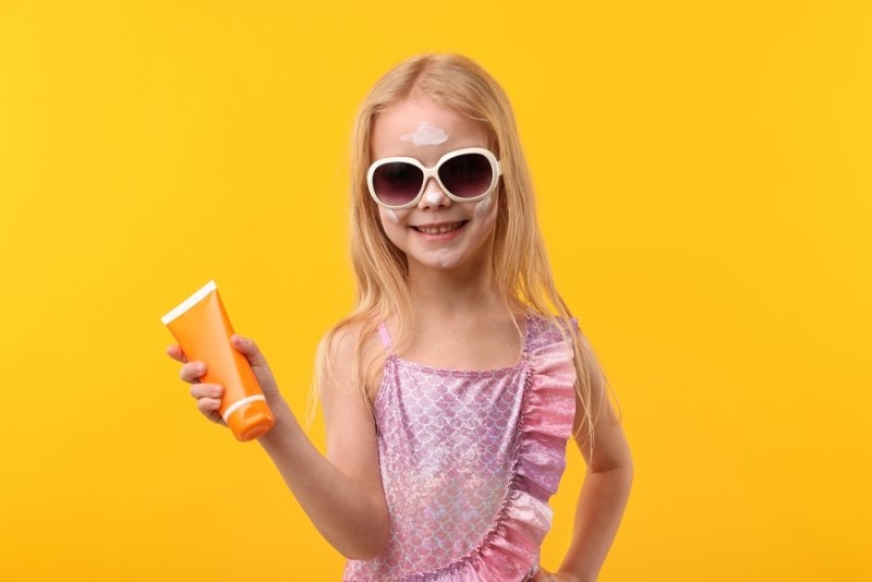 Smiling little girl with tube of sun protection cream against yellow background.
