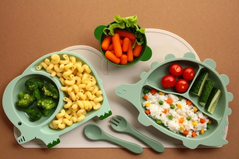 Child-friendly plates containing pasta, rice, broccoli, carrots, tomatoes, and cucumbers arranged with children's utensils.