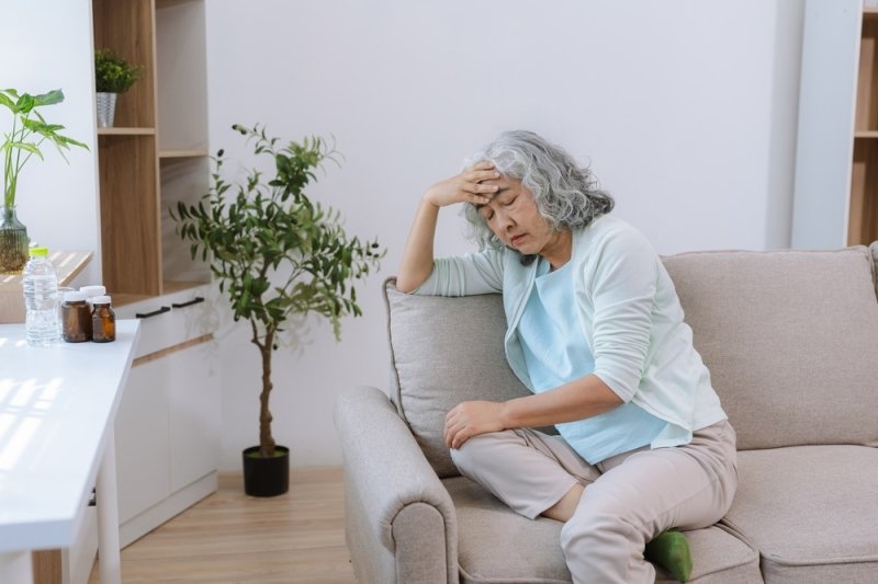 Older woman sitting on a sofa with her eyes closed and one hand on her forehead, appearing uncomfortable in a bright living room.