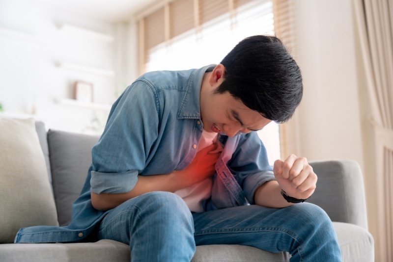 Young man sitting on a sofa and clutching his chest, with a red visual effect highlighting the heart area to represent chest discomfort.