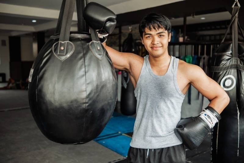A smiling boxer stands confidently beside a punching bag, wearing training gloves in a boxing gym.