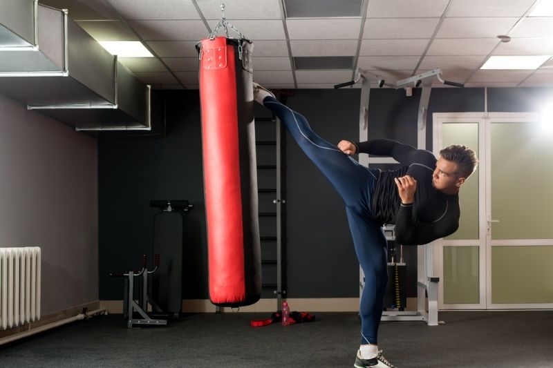 Man performing a high kick on a red punching bag during a martial arts workout.