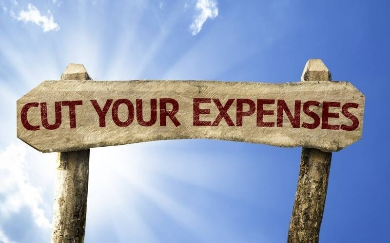A rustic wooden sign displaying “CUT YOUR EXPENSES” against a bright blue sky.