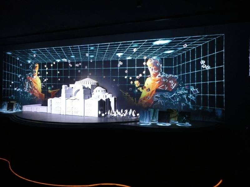 Holographic digital art display featuring architectural models and figures in a dark exhibition space.