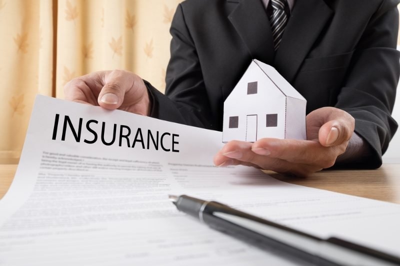 A person in a business suit holding an insurance document and a small paper house.