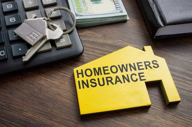 A yellow house-shaped sign labeled “HOMEOWNERS INSURANCE,” alongside house keys, a calculator, cash, and documents on a wooden desk.