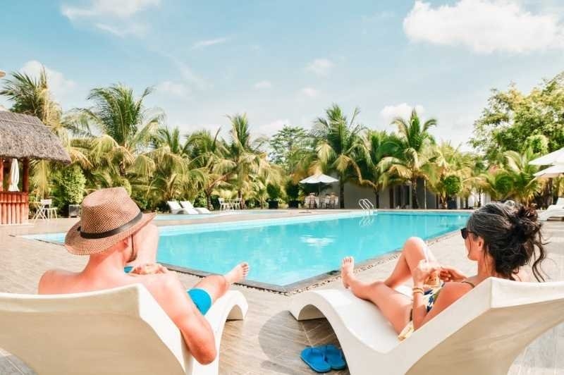 Couple lay on sunbeds enjoy sunbathing drink cold beverage on all-inclusive vacation holidays at tropical resort with swimming pool surrounded by coconut palm trees by coast on sunny hot tropical day.
