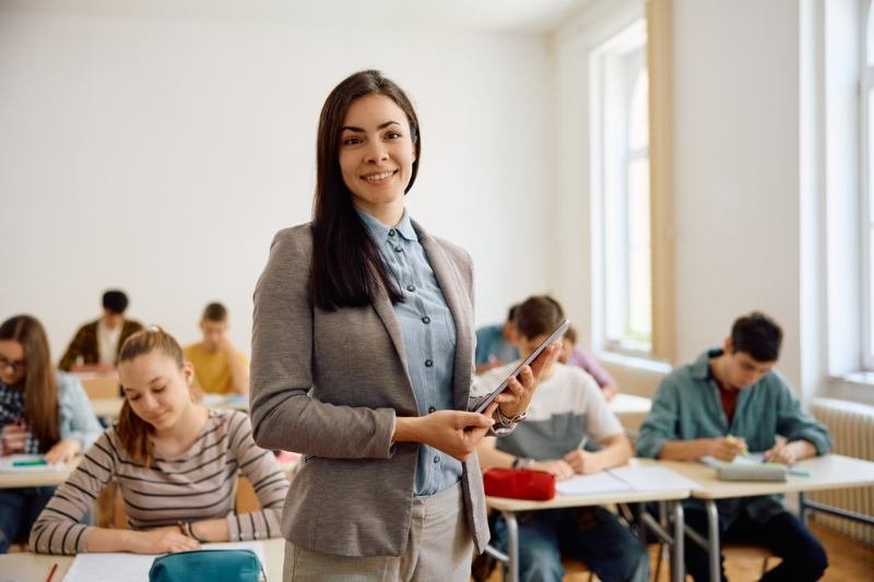 A female teacher stands at the front of a classroom holding a tablet or clipboard while students work at their desks in the background.