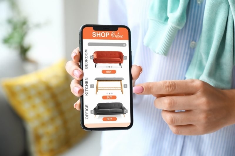 A person holding a smartphone displaying an online furniture store with products and “BUY” buttons.
