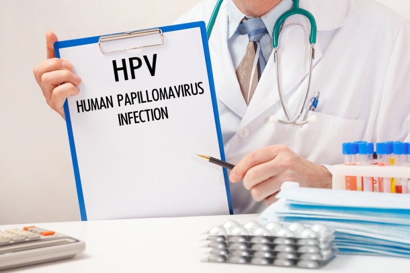 Doctor holding a clipboard labeled “HPV Human Papillomavirus Infection” in a medical setting with a stethoscope, test tubes, and medication in the foreground.
