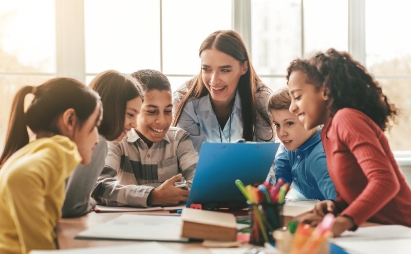 Teacher working with a diverse group of children gathered around a tablet in a classroom.