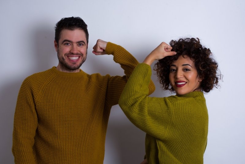 Smiling man and woman wearing colorful sweaters while flexing their arms to symbolize individuality and self-expression.