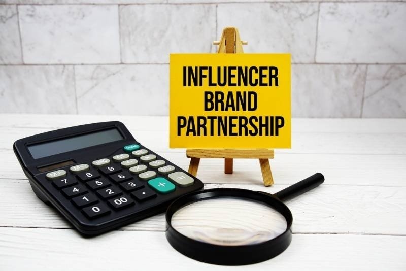 A yellow sign on a small easel reads “INFLUENCER BRAND PARTNERSHIP,” surrounded by a calculator and magnifying glass on a wooden surface.