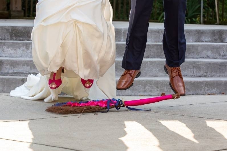 Bride and groom jumping over a decorated ceremonial broom during an outdoor wedding.