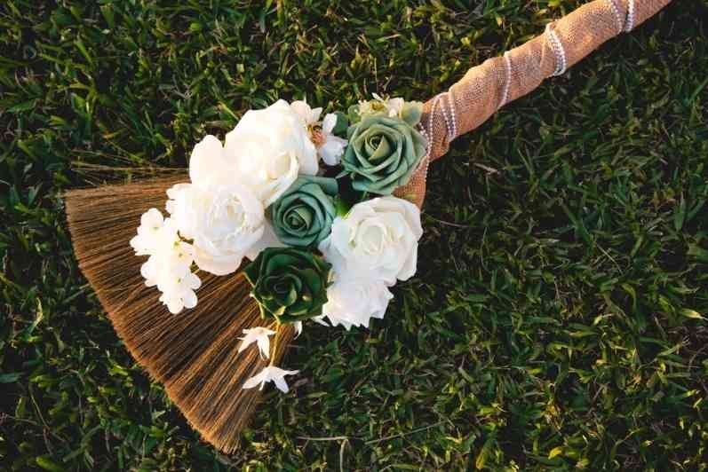 Traditional wedding broom decorated with white and green flowers resting on grass.