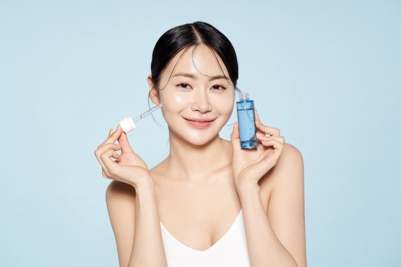 Woman holding a blue skincare serum bottle and dropper beside her face against a light blue background, illustrating a Korean skincare routine.