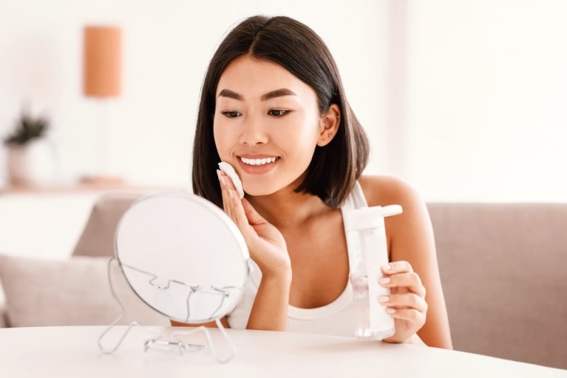 Woman applying facial toner with a cotton pad while looking into a tabletop mirror, holding a skincare bottle in a bright bathroom setting.
