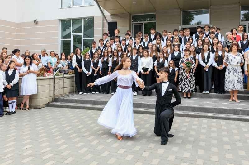 A large group of students, teachers, and families gather outside a school building while two students perform a formal dance in front of the crowd.