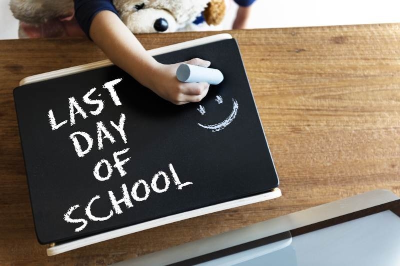 A child writes “LAST DAY OF SCHOOL” in large white chalk letters on a blackboard, with a smiley face beside the message.