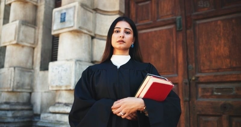 Professional female lawyer in a black legal robe holding law books outside a courthouse.