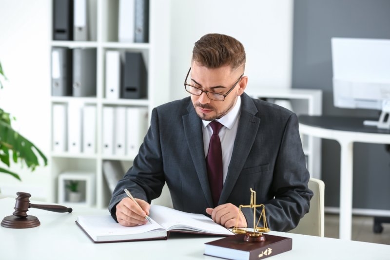 A lawyer in a suit writes in a legal book at a desk, with a gavel and scales of justice nearby.