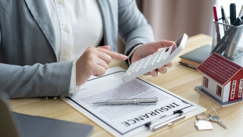 Person using a calculator beside insurance documents, a model house, and keys.
